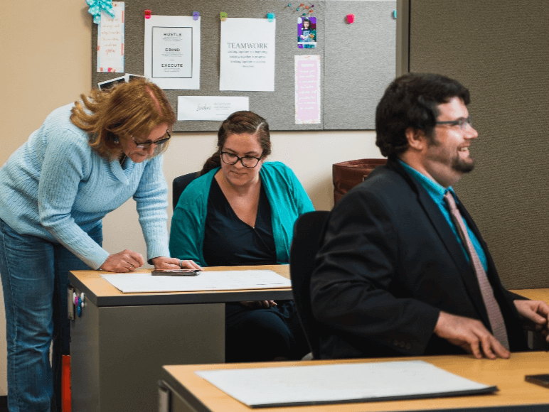 Two Answering Service for Directors' employees having a discussion at a desk, while another smiles at his desk in the foreground