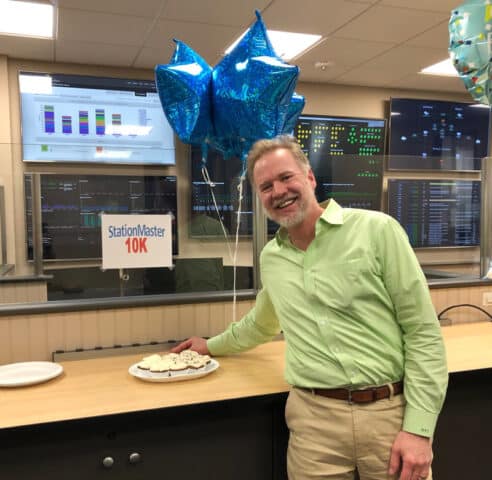 ASD employee standing in front of balloons and a plate of cupcakes in the office to celebrate the 10,000th update to the answering service system.